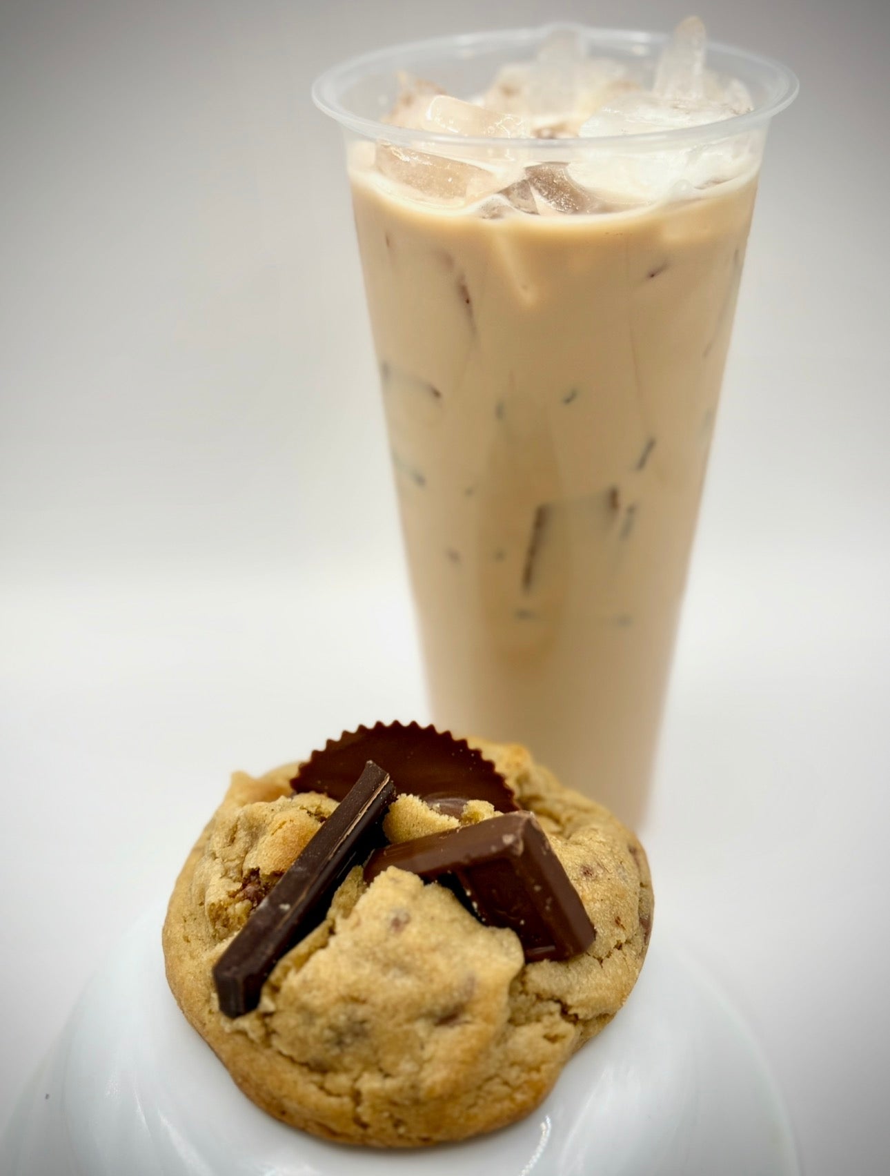 Peanut butter cookie with chocolate pieces next to a glass of iced coffee on a white background