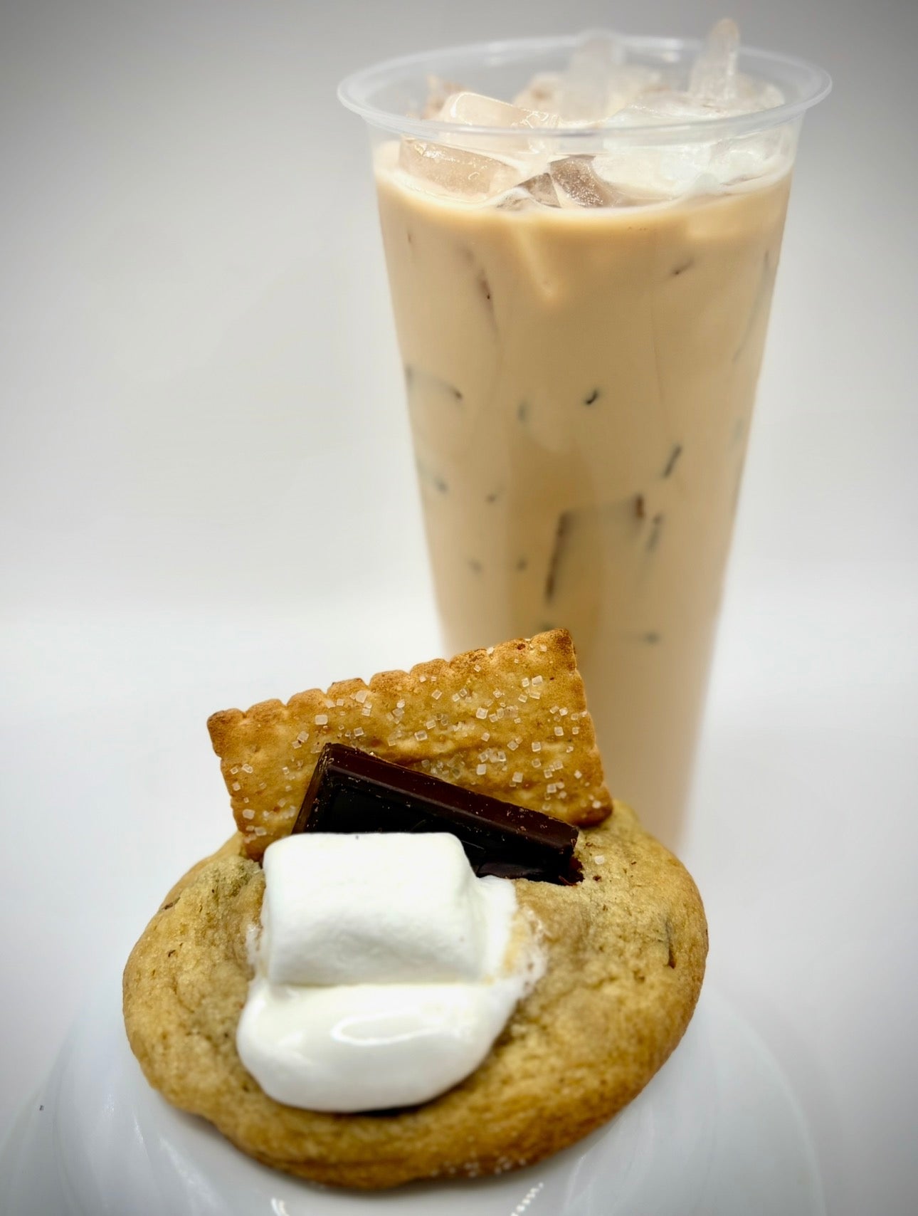 Cookie with marshmallows and a chocolate bar next to an iced coffee drink on a white background